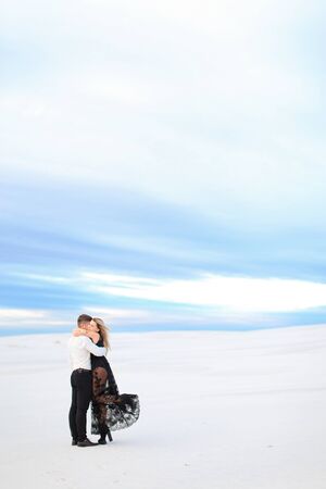 Young happy husband in white shirt hugging wife wearing black transparent dress in winter steppe. Concept of winter photo session, relationship and love, happy couple.の写真素材