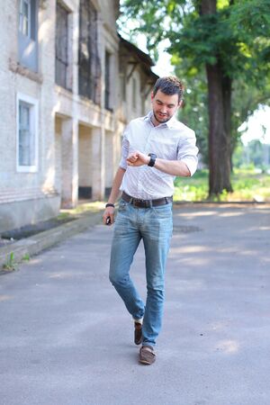 Bearded young men man guy goes, walking along road and looking at watch on arm. Young man dressed in white shirt with red stripe, blue jeans and brown loafers. Concept of business man waiting for meeting, posing in front of camera, casual style.の写真素材