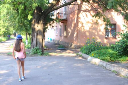 Girl child stands with his back to the photographer, goes, directed towards the house, walking. Girl dressed in pink shirt, Printed shorts, dark blue hat with a brim and blue sandals. Concept of summer holidays, childhood, walk.の写真素材