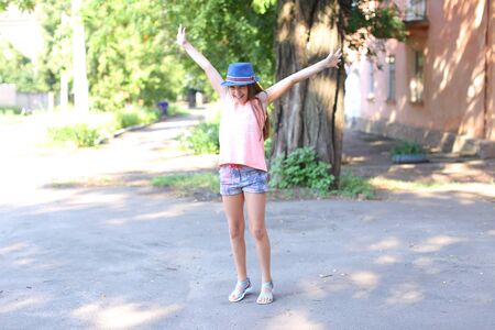 Pretty cute female girl child jumping, fun, fooling around and laughing in yard on street summer day. Girl dressed in pink shirt, printed shorts, dark blue hat with brim and blue sandals. Concept of fun, children happiness, childhood, summer holidays.の写真素材