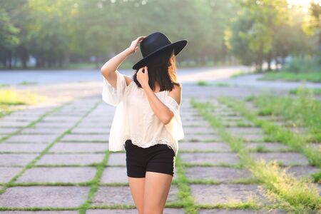 female nature keeps his hat summer sun sunset looks pleased. azitka woman black hair white shirt black shorts dancing around the trees looking at the camera. portrait. spinningの写真素材