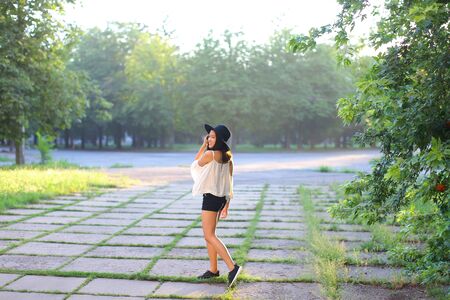 female nature keeps his hat summer sun sunset looks pleased. azitka woman black hair white shirt black shorts dancing around the trees looking at the camera. portrait. spinningの写真素材