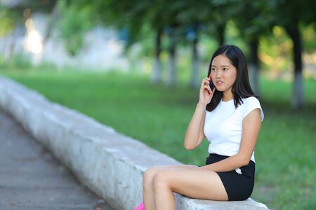 Girl female teenager with black hair in a bob white east Asian woman green park with a backpack background. Lady holds, dials, push your finger, smiling, looking into the camera, talking on the phone happy.の写真素材