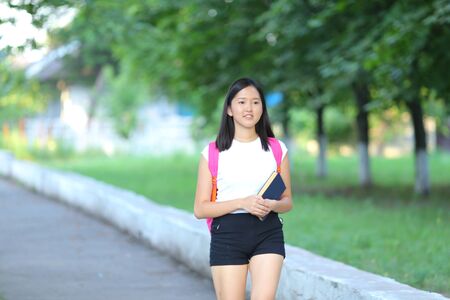 Girl female teenager with black hair in a bob white east Asian woman green park with a backpack background. Lady walking, goes, walk away, look at the camera.の写真素材