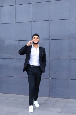 Entrepreneur man male looking at camera and smiling talks on mobile smart phone. Young handsome businessman arab muslims in business center wearing dressed in black elegant suit on building background.の写真素材