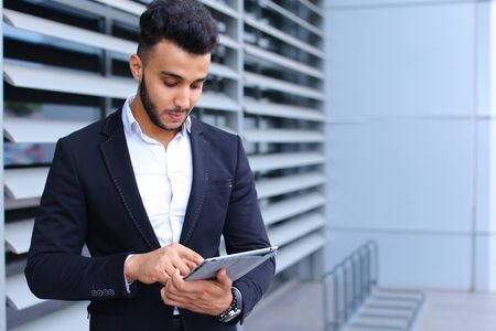 Young handsome businessman Arab Muslims is in the business center in black elegant dress on building background. Businessman man male looking waiting concept and smiling.の写真素材