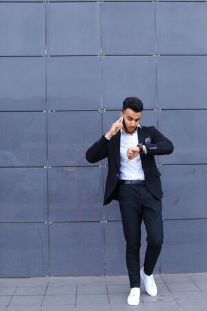 Entrepreneur man male looking at camera and smiling talks on mobile smart phone. Young handsome businessman arab muslims in business center wearing dressed in black elegant suit on building background.の写真素材