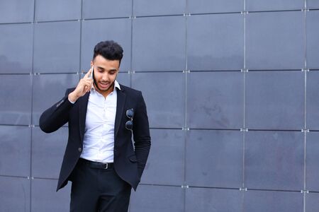 Entrepreneur man male looking at camera and smiling talks on mobile smart phone. Young handsome businessman arab muslims in business center wearing dressed in black elegant suit on building background.の写真素材