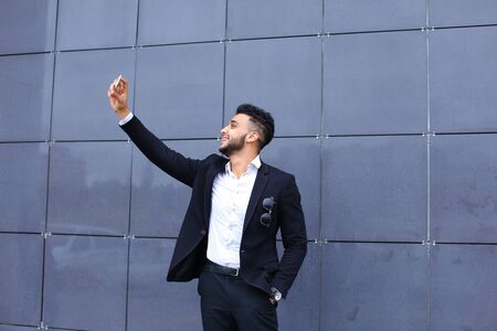 Entrepreneur takes makes selfie man male looking at camera and smiling talks on mobile smart phone. Young handsome businessman arab muslims in business center wearing dressed in black elegant suit on building background.の写真素材