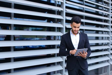 Young handsome businessman Arab Muslims is in the business center in black elegant dress on building background. Businessman man male looking waiting concept and smiling.の写真素材