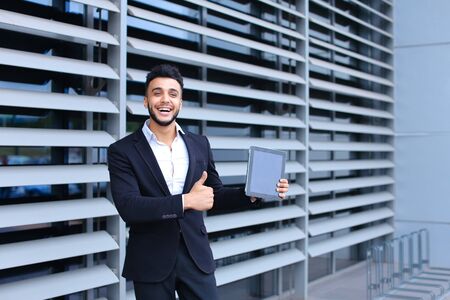 Young handsome businessman Arab Muslims is in the business center in black elegant dress on building background. Businessman man male looking waiting concept and smiling.の写真素材