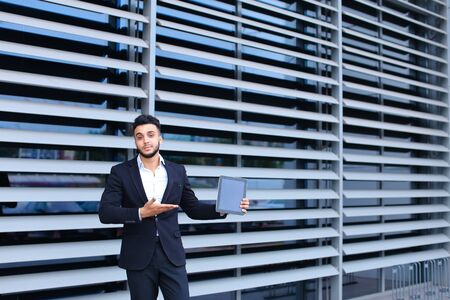Young handsome businessman Arab Muslims is in the business center in black elegant dress on building background. Businessman man male looking waiting concept and smiling.の写真素材