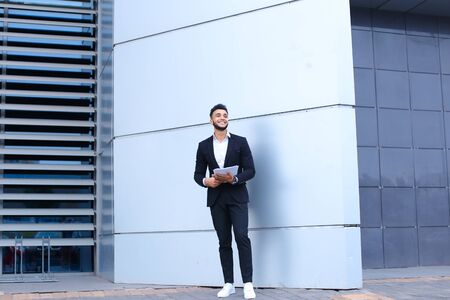 Young Arab male man businessman smiling and holds securities or documents, stands near gray wall of business center.の写真素材
