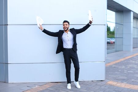 Young beautiful male guy arab businessman student laughing, smiling, rejoices success, holds papers or documents, stands in full growth directly into camera against background of gray wall of large shopping business center.の写真素材
