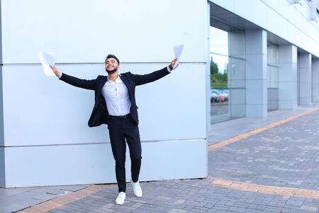 Young beautiful male guy arab businessman student laughing, smiling, rejoices success, holds papers or documents, stands in full growth directly into camera against background of gray wall of large shopping business center.の写真素材