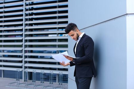 Perspective young adult businessman virile asian guy holds and looks, considers, reads and sorts out papers or documents, stands near gray wall of business center.の写真素材