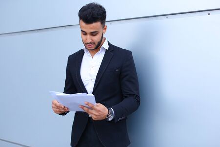 Young successful businessman arab man holds documents, securities, smiling little smile and thinks near gray wall of business center. Muscular bearded man dressed in classic suit, black pants, jacket , white shirt. Concept of securities, Business Meeting, busy Dealer.の写真素材