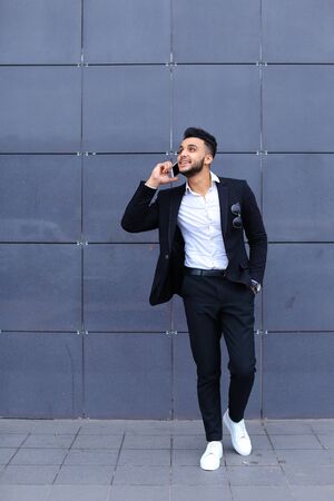 Entrepreneur man male looking at camera and smiling talks on mobile smart phone. Young handsome businessman arab muslims in business center wearing dressed in black elegant suit on building background.の写真素材