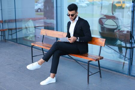 Entrepreneur smiling man puts on sunglasses uses tablet and looks at. Young handsome businessman arab muslims in business center wearing dressed in black elegant suit on building background.の写真素材