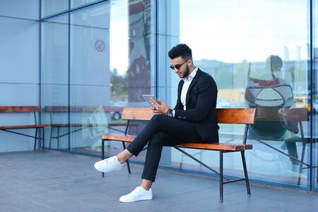 Entrepreneur smiling man puts on sunglasses uses tablet and looks at. Young handsome businessman arab muslims in business center wearing dressed in black elegant suit on building background.の写真素材