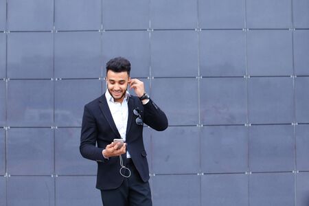 Entrepreneur takes makes selfie man male looking at camera and smiling talks on mobile smart phone. Young handsome businessman arab muslims in business center wearing dressed in black elegant suit on building background.の写真素材