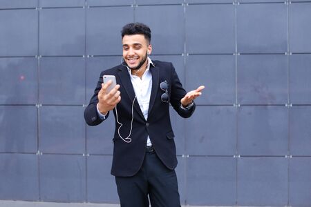 Entrepreneur takes makes selfie man male looking at camera and smiling talks on mobile smart phone. Young handsome businessman arab muslims in business center wearing dressed in black elegant suit on building background.の写真素材