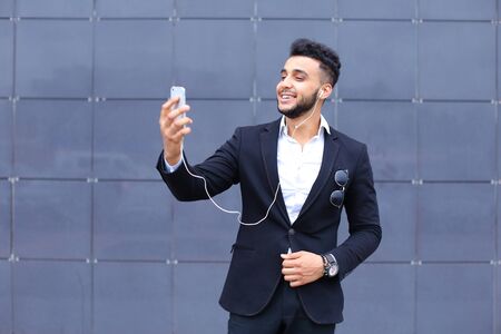 Entrepreneur takes makes selfie man male looking at camera and smiling talks on mobile smart phone. Young handsome businessman arab muslims in business center wearing dressed in black elegant suit on building background.の写真素材