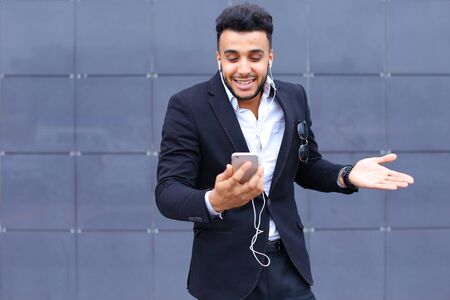 Entrepreneur takes makes selfie man male looking at camera and smiling talks on mobile smart phone. Young handsome businessman arab muslims in business center wearing dressed in black elegant suit on building background.の写真素材