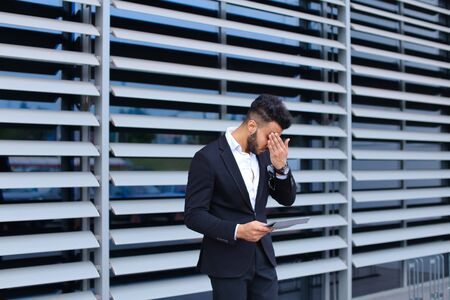 Handsome young male businessman man holds and uses tablet for communication, business, near gray wall with shutters of business center.の写真素材