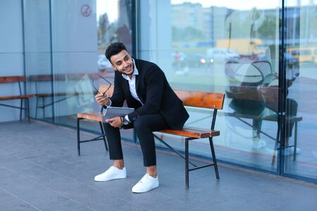 Entrepreneur smiling man puts on sunglasses uses tablet and looks at. Young handsome businessman arab muslims in business center wearing dressed in black elegant suit on building background.の写真素材