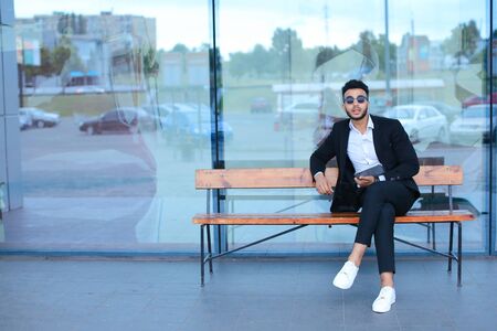 Entrepreneur smiling man puts on sunglasses uses tablet and looks at. Young handsome businessman arab muslims in business center wearing dressed in black elegant suit on building background.の写真素材