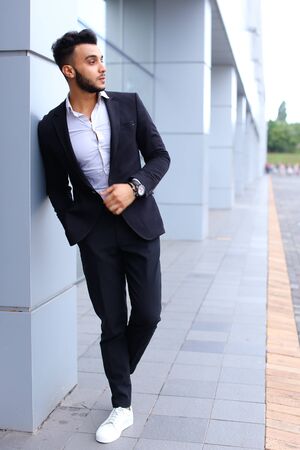 Young handsome businessman Arab Muslims is in the business center in black elegant dress on building background. Businessman man male looking at time walking goes and smiling.の写真素材