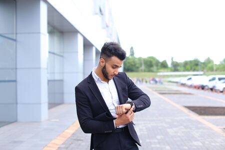 Young handsome businessman Arab Muslims is in the business center in black elegant dress on building background. Businessman man male looking at time walking goes and smiling.の写真素材