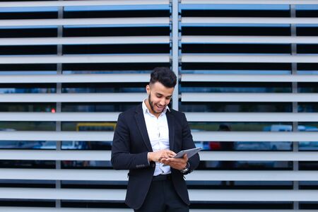 Entrepreneur uses tablet for online order shopping with debit credit card looking smiling at the camera. Young handsome businessman arab muslims in business center wearing dressed in black elegant suit on building background.の写真素材