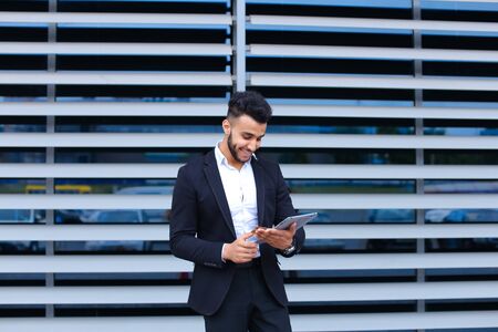 Entrepreneur uses tablet for online order shopping with debit credit card looking smiling at the camera. Young handsome businessman arab muslims in business center wearing dressed in black elegant suit on building background.の写真素材