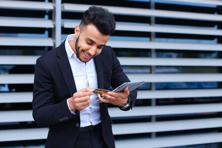 Entrepreneur uses tablet for online order shopping with debit credit card looking smiling at the camera. Young handsome businessman arab muslims in business center wearing dressed in black elegant suit on building background.の写真素材