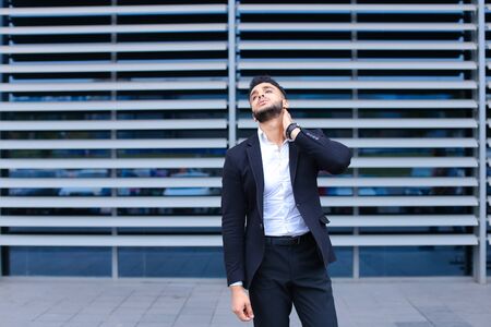 Entrepreneur shows neck pain tired looking at the camera. Young handsome businessman arab muslims in business center wearing dressed in black elegant suit on building background.の写真素材