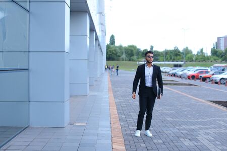 Young handsome businessman Arab Muslims is in the business center in black elegant dress on building background. Businessman man male looking at time walking goes and smiling.の写真素材
