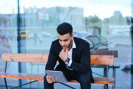 Entrepreneur smiling man puts on sunglasses uses tablet and looks at. Young handsome businessman arab muslims in business center wearing dressed in black elegant suit on building background.の写真素材