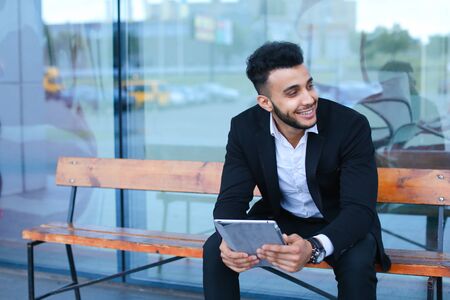 Entrepreneur smiling man puts on sunglasses uses tablet and looks at. Young handsome businessman arab muslims in business center wearing dressed in black elegant suit on building background.の写真素材