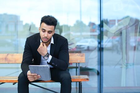 Entrepreneur smiling man puts on sunglasses uses tablet and looks at. Young handsome businessman arab muslims in business center wearing dressed in black elegant suit on building background.の写真素材