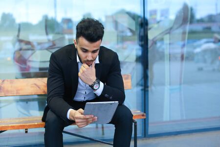Entrepreneur smiling man puts on sunglasses uses tablet and looks at. Young handsome businessman arab muslims in business center wearing dressed in black elegant suit on building background.の写真素材