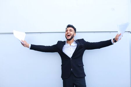One handsome young adult arabian guy entrepreneur student smiling broadly and holds paper on background of gray wall.Muscular bearded man dressed in classic suit, black pants, jacket , white shirt, on hand is watch.Concept of successful businessman, joy of good bargain, securities, Business Meeting, busy Dealer.の写真素材