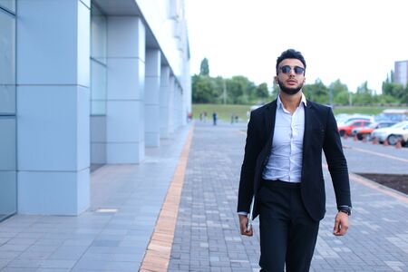 Young handsome businessman Arab Muslims is in the business center in black elegant dress on building background. Businessman man male looking at time walking goes and smiling.の写真素材