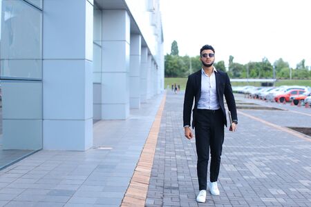 Young handsome businessman Arab Muslims is in the business center in black elegant dress on building background. Businessman man male looking at time walking goes and smiling.の写真素材