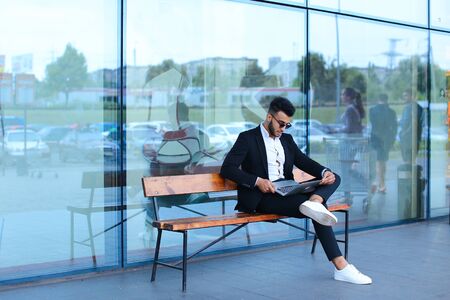 Entrepreneur uses laptop wearing sunglasses. Young handsome businessman arab muslims in business center wearing dressed in black elegant suit on building background.の写真素材