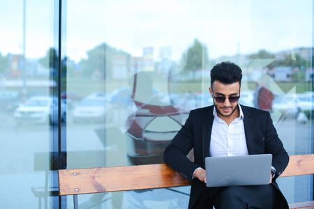 Entrepreneur uses laptop wearing sunglasses. Young handsome businessman arab muslims in business center wearing dressed in black elegant suit on building background.の写真素材