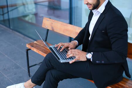 Entrepreneur uses laptop wearing sunglasses. Young handsome businessman arab muslims in business center wearing dressed in black elegant suit on building background.の写真素材