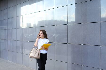 Serious young adult business female woman talks on phone, negotiating and looks aside, holds documents in yellow folder on background of gray wall of shopping business center.の写真素材