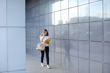 Serious young adult business woman walks and talks on phone, solves problems, holds and looks at documents in yellow folder on background of gray wall of shopping business center.の写真素材